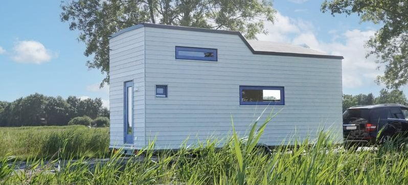 Weiß-blaues Tiny House auf Anhänger vor Wiese und Bäumen